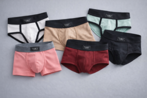 TAMZ men’s underwear collection featuring solid color briefs and trunks in beige, pink, burgundy, mint, white, and black laid flat on a soft gray fabric background with visible logo waistbands.